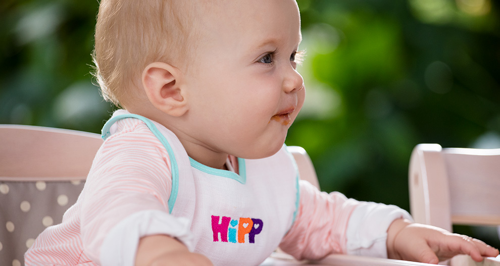 Syöttötuolissa istuva vauva, jolla on ruokalappu, jossa lukee "HIPP", katsoo sivulle.
